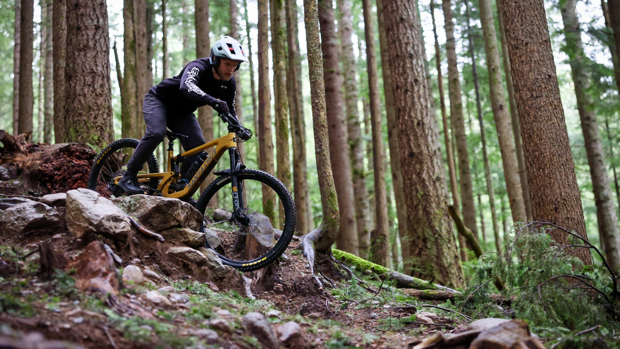 Mountain biker riding a Santa Cruz Bronson down steep, rocky, and root trail in the Pacific Northwest woods.