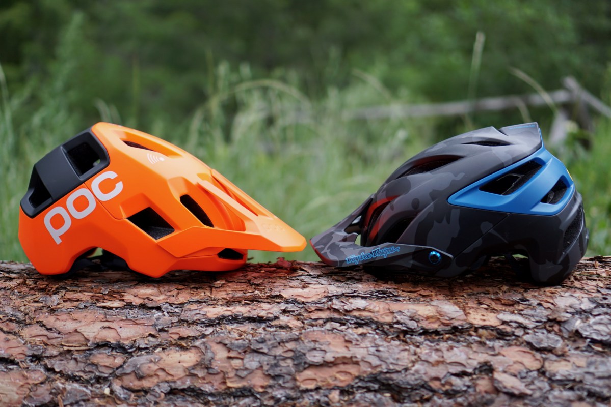 Black and blue Troy Lee Designs A3 vs. orange and black POC Kortal Race MIPS mountain bike helmets sitting on a log.